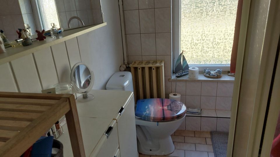 Kleines, gefliestes Badezimmer mit WC, weißem Unterschrank und Wandregal mit Spiegel. Daneben ein großes Fenster mit Milchglas, darunter eine geflieste Ablagefläche sowie ein Heizkörper in der Ecke.