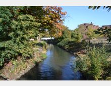 Ein Fluss fließt durch eine von Bäumen und Sträuchern gesäumte Landschaft mit einem Backsteingebäude im Hintergrund. Im oberen Bildbereich ist eine weiße Brücke sichtbar, die den Fluss überquert.