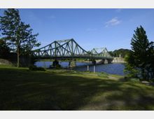 Grüne Stahlbrücke mit Bögen und Gitterträgern, die über einen Fluss führt, umgeben von einer Parklandschaft mit Bäumen, Bänken und Rasenflächen unter blauem Himmel.
