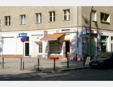 Kleiner Eckladen in einem mehrstöckigen Wohnhaus mit heller Fassade, mehreren Schaufenstern und einer orangefarbenen Markise. Vor dem Laden stehen Schilder und Plakate, auf der Straße befinden sich Verkehrsschilder sowie geparkte Autos.