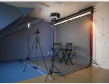Fotostudio mit einem grauen Hintergrund, zwei schwarzen Regiestühlen und Beleuchtungsausrüstung einschließlich Stativen und Softboxen. Der Boden ist zweifarbig, und die Wand hat blaue und beige Bereiche.