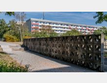 Außenbereich mit einer Betonmauer aus ornamentierten Blöcken, die einen Weg mit Pflastersteinen begrenzt. Im Hintergrund ist ein mehrstöckiges Wohngebäude mit Balkonen und roten Akzenten, umgeben von Bäumen und Sträuchern zu sehen.