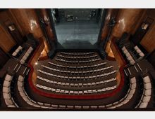 Innenansicht eines Theatersaals mit halbrunden Sitzreihen, die auf die Bühne ausgerichtet sind. Die Wände sind aus Holzverkleidung, und der Hintergrund zeigt die leere Bühne mit technischem Equipment.