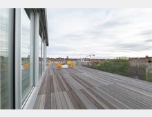Weitläufige Dachterrasse mit Holzdielen, gelben Liegestühlen und einem kleinen weißen Tisch, umgeben von einer Glasbrüstung; im Hintergrund ist eine Stadtkulisse mit Baukränen und Gebäuden zu sehen. Links spiegelt sich eine Glasfassade eines angrenzenden Gebäudes.
