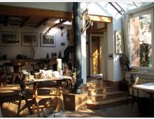 Künstleratelier mit großen Fenstern, die Tageslicht hereinlassen. Im Vordergrund steht ein Tisch mit Malutensilien, im Hintergrund sind Holzstühle, ein runder Tisch, Bilder an der Wand sowie ein Kaminofen zu sehen.