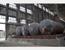 Große, verrostete Metalltanks stehen auf einem Podest in einer verlassenen Fabrikhalle mit schwarzen Ziegelwänden und hohen, teils zerbrochenen Fensterreihen. Die Halle ist leer, und der Boden besteht aus Schutt und Staub.