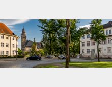 Straßenszene mit Kopfsteinpflaster, flankiert von einem mehrstöckigen Wohngebäude links und einem hellen, historischen Gebäude mit rot gerahmten Fenstern rechts. Im Hintergrund ist ein Kirchturm mit Uhr und grüner Baumvegetation sichtbar.