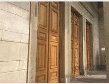 Sandfarbenes Gebäude mit großen, verzierten Holztüren und mehreren Säulen, die einen Eingangsbereich mit hellen Steinwänden einrahmen. Der Boden besteht aus grauen Steinplatten.