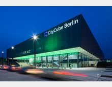 Außenansicht des CityCube Berlin bei Nacht, einem modernen Veranstaltungszentrum mit klaren Glas- und Metallfassaden. Das Gebäude ist von grüner Beleuchtung erhellt, darunter eine beleuchtete Eingangshalle mit großen Glasfronten und automatischen Türen.
