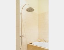 Eine Badezimmerecke mit cremefarbenen Fliesen, einer runden Regendusche aus Edelstahl und einer eingelassenen Badewanne mit beige gefliestem Rand. Im Vordergrund stehen eine Bürste und eine Kerze auf dem Wannenrand.