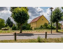 Gelbes, zweistöckiges Wohnhaus mit rotem Satteldach, umgeben von Bäumen, Sträuchern und einer offenen Grünfläche. Im Vordergrund führen ein Bürgersteig und eine Straße vorbei, flankiert von Laternen und einzelnen Bäumen.