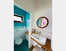 Kleines Badezimmer mit türkisfarben gestrichenen Wänden und weißer Fliesenverkleidung, links mit wandhängendem WC und kleinem Fenster. Rechts ein Waschbecken auf Holzplatte mit rundem Wandspiegel darüber.