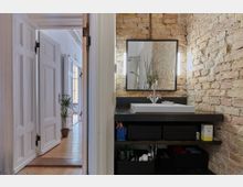 Badezimmer mit einem Waschbecken auf einer dunklen Ablage, einem großen rechteckigen Spiegel und sichtbarer Backsteinwand. Im angrenzenden Flur sind helle Holzböden, weiße Türen und eine Topfpflanze zu sehen.