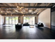 Industrie-inspirierter Innenraum mit schwarzem Sofa-Set und Glastischen auf einem dunklen Boden. Die Decke zeigt freiliegende Betonträger und Lüftungsrohre, während große Glasfenster mit Vorhängen Blick auf eine Straße mit parkenden Autos ermöglichen.