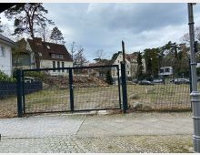 Ein leeres, umzäuntes Grundstück in einer Wohngegend mit gepflastertem Gehweg im Vordergrund; im Hintergrund stehen Häuser mit mehreren Stockwerken und Satteldächern sowie Bäume unter einem bewölkten Himmel.