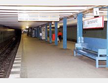 Untergrundbahnhof mit blau gestrichenen Säulen, einem leeren Gleis links und einem Bahnsteig mit Sitzbänken und Schildern. Im Hintergrund ist das Stationsschild „Spittelmarkt“ sichtbar, sowie eine Person, die sich entfernt.