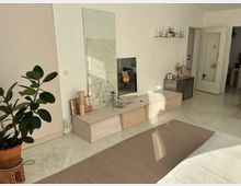 Helles Wohnzimmer mit minimalistischem Design. Im Vordergrund steht eine grüne Zimmerpflanze in einem Terrakottatopf, dahinter ein niedriger Holzschrank mit Dekorationen und einem großen Spiegel an der weißen Wand. Rechts sind eine Tür und ein Regal mit Geschirr zu sehen.