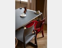 Ein moderner Innensitzbereich mit einem grauen Tisch, dekoriert mit kleinen Pflanzen, Keramikgefäßen und Tellern. Zwei schlichte Kunststoffstühle in Rot und Grau stehen auf einem hellen Parkettboden, während Tageslicht durch ein Fenster mit weißen Vorhängen einfällt.