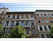 Fassade eines alten, teils heruntergekommenen Gebäudes mit Graffiti und protestartigen Schriftzügen, darunter groß „Kapitalismus zerstört“ und „tötet“. Daneben ein renoviertes Gebäude mit klarer Backstein- und Putzarchitektur. Im Vordergrund sind Bäume und ein Straßenlaternenmast sichtbar.