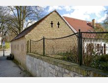 Backsteingebäude mit schrägem Dach, zwei kreuzförmigen Öffnungen in der Giebelfassade und angrenzendem Metallzaun auf einer Steinmauer; im Hintergrund ein weiteres Haus mit rot gedecktem Dach und weißer Fassade.