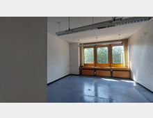 Ein leerer Raum mit hellen Wänden, einem blauen Boden und großen Fenstern mit orangefarbenen Rahmen, die auf eine grüne Außenszenerie blicken. Von der Decke hängen längliche LED-Leuchten, und unter den Fenstern befinden sich Paneele mit Steckdosen.