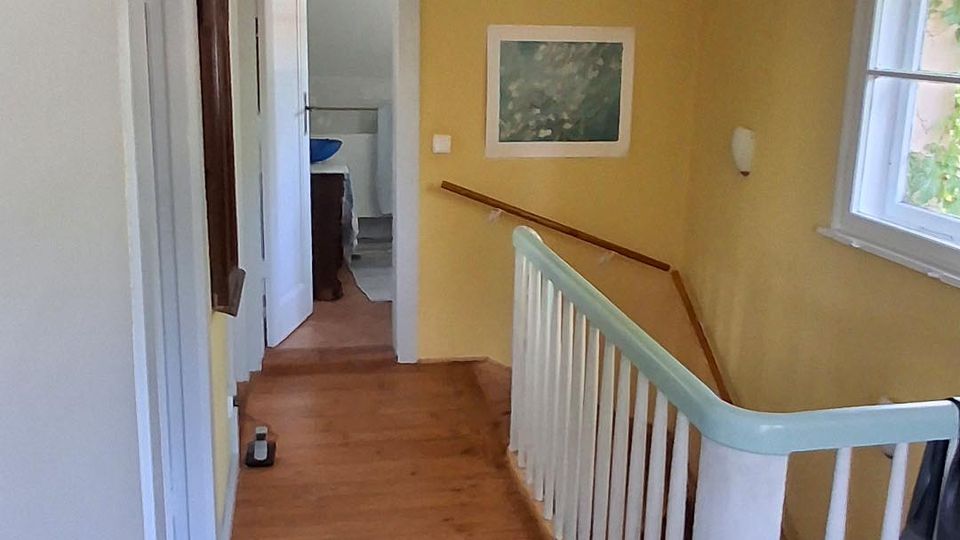 Schmaler Flur in einem Wohnhaus mit gelb gestrichenen Wänden, Holzdielenboden und weißer Decke mit einfacher Hängelampe. Rechts führt ein Treppenabgang mit weißem Geländer nach unten, daneben ein Fenster; am Ende des Flurs ist eine offene Tür zu einem angrenzenden Raum sichtbar.