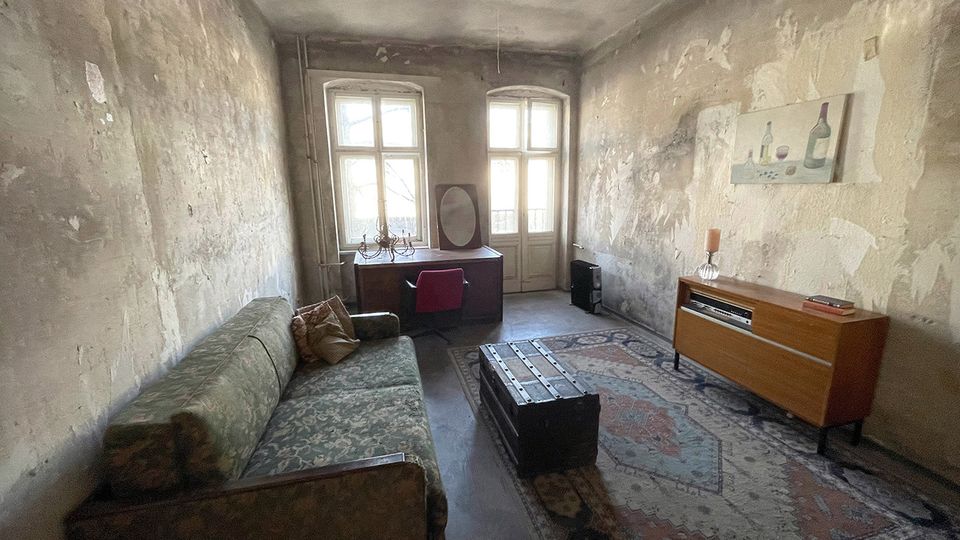 Langer, schmaler Wohnraum mit abgenutzten, teilweise abgeplatzten Wänden und zwei hohen Fenstern am Ende, die viel Tageslicht hereinlassen. Im Raum stehen ein altes Sofa, ein Schreibtisch mit Stuhl, ein großer Teppich, ein trunkähnlicher Couchtisch sowie ein Sideboard an der rechten Wand.