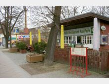 Außenansicht eines kleinen Cafés mit der Aufschrift „Café-Restaurant Am Park“ in einer urbanen Umgebung. Im Vordergrund befinden sich Sitzbänke und Tische im Freien, umgeben von Pflanzen und Bäumen; im Hintergrund ist ein Schild der U-Bahn-Station und umliegende Gebäude zu sehen.