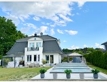 Modernes Einfamilienhaus mit weißer Fassade und dunklem Mansarddach, davor eine große Terrasse und ein langes, flaches Glasdach über einem Pool. Der Garten ist von Rasen, Hecken und Bäumen umgeben, mit Pavillons und Sitzmöbeln unter blauem Himmel mit Wolken.
