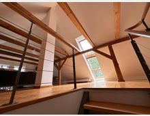Moderne Dachgeschosswohnung mit sichtbaren Holzbalken, hellem Laminatboden und schrägen Wänden. Ein großes Dachfenster lässt viel Licht herein, und eine Metallgeländerkonstruktion trennt den Treppenaufgang vom Wohnbereich.