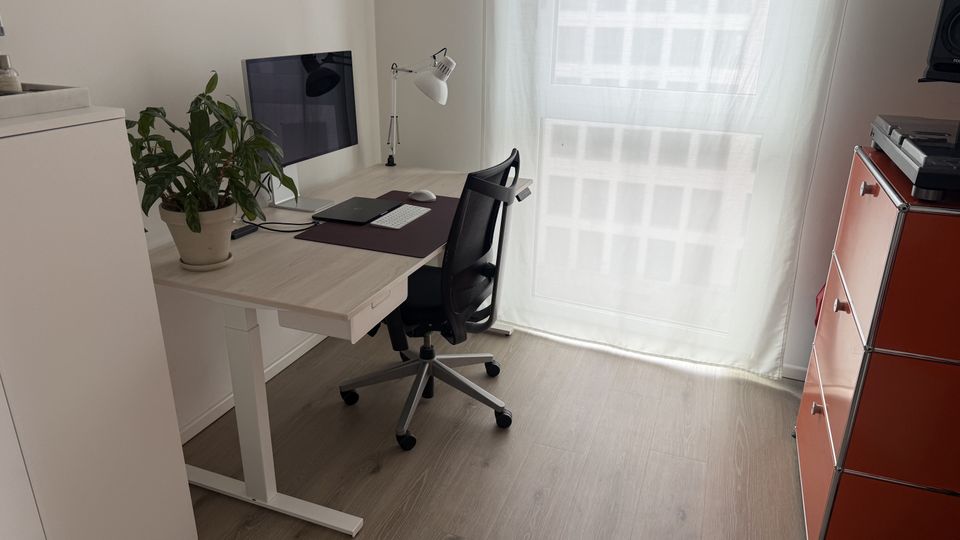 Helles, minimalistisches Homeoffice mit höhenverstellbarem Schreibtisch, Monitor, Schreibtischlampe und Drehstuhl auf grauem Holzfußboden. Ein großes Fenster mit transparentem Vorhang lässt Tageslicht herein; daneben steht ein rotes Schubladenregal.
