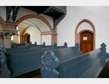 Innenraum einer historischen Kirche mit dunkelblau gestrichenen Holzbankreihen, verzierten Armlehnen und rötlich-braunen Backsteinbögen. Im Hintergrund befindet sich eine Holztür mit einem Schild "Kein Durchgang".