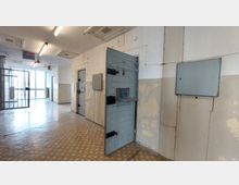 Ein Flur in einem Gefängnis oder einer ähnlichen Einrichtung mit mehreren grauen Zellentüren, von denen eine geöffnet ist. Die Wände sind teilweise verputzt und abgeblättert, der Boden ist mit einem abgenutzten Linoleumbelag versehen, und am Ende des Flurs befindet sich ein vergittertes Fenster mit einem Gittertor.