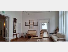 Heller Wohnraum im klassischen Stil mit weißen Wänden, Holzfußboden und großen Fenstern mit Vorhängen. Zu sehen sind Sitzmöbel, ein Wandspiegel, gerahmte Zeichnungen sowie breite Türdurchgänge in angrenzende Räume, darunter ein Raum mit Kronleuchter.