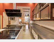 Moderne Gewerbeküche mit Edelstahl-Arbeitsflächen, weißen Fliesenwänden und einer schwarzen Dunstabzugshaube. Im Hintergrund befindet sich ein angrenzender Essbereich mit Holzstühlen und großen Fenstern, die Tageslicht hereinlassen.