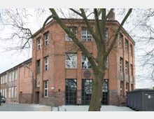 Backsteinbau im industriellen Stil mit großen Fenstern und einem mehrstöckigen Hauptgebäude, flankiert von einem angrenzenden Seitenflügel. Ein großer, blattloser Baum steht im Vordergrund, und der Boden davor ist gepflastert.