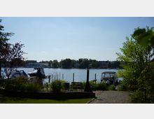 Uferansicht mit Blick auf einen Fluss oder See, im Vordergrund eine gepflasterte Fläche, Gartenmöbel und Pflanzen, links und rechts Boote an Stegen. Im Hintergrund befinden sich Gebäude und Bäume am gegenüberliegenden Ufer.