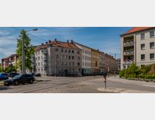 Straßenecke in einem Wohngebiet mit mehrstöckigen Wohnhäusern, teils in pastellfarbenem Putz. Kopfsteinpflaster und Straßenbahnschienen prägen die Straße, während geparkte Autos und begrünte Bereiche den Straßenrand säumen.