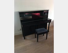 Schwarzes Klavier mit glänzender Oberfläche, positioniert vor einer weißen Wand auf einem hellen Holzboden, daneben ein dazugehöriger schwarzer Klavierhocker.