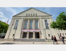 Außenansicht eines Kultur- und Festspielhauses im neoklassizistischen Stil mit einer symmetrischen Fassade, großen rechteckigen Fenstern und vier glatten Säulen. Vor dem Gebäude stehen Flaggenmasten und einige Personen unterhalten sich.