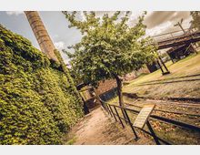 Industriegelände mit historischem Charakter, bestehend aus einem mit Efeu bewachsenen Gebäude, einem hohen Schornstein, einem Baum und alten Gleisanlagen. Im Vordergrund befindet sich ein Geländer und ein Schild, auf dem offenbar Informationen angegeben sind.