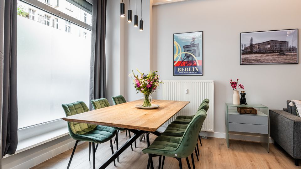 Heller, modern eingerichteter Essbereich mit großem Holztisch und mehreren grünen Polsterstühlen auf hellem Holzboden. Links ein bodentiefes Fenster mit Vorhängen, darüber hängende Pendelleuchten; an der Wand Heizkörper und gerahmte Bilder sowie ein kleines Sideboard.