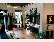 Ein stilvolles Zimmer mit Holzdielenboden, Pflanzen in Töpfen und einer Kombination aus modernen und klassischen Elementen. Ein dunkles Holzmöbelstück mit Regalen steht an der Wand, darüber hängen Gemälde, während Licht durch die Fenster fällt und den Raum erhellt.