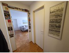 Flur in einer Wohnung mit hellem Parkettboden und weißen Wänden. Links hängt eine Sammlung von Postkarten um die Türzarge, die in ein Wohnzimmer mit einer grauen Couch und einem Bild an der Wand führt. Rechts an der Wand ist ein gerahmtes Poster mit einer Illustration von Berlin zu sehen.