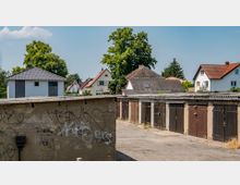 Reihen von alten Garagen mit Holztüren und Graffiti auf einer Betonwand im Vordergrund; im Hintergrund sind Einfamilienhäuser mit Satteldächern sowie große Bäume zu sehen.