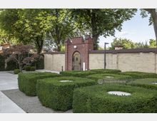 Eine gepflegte Gartenanlage mit rechteckigen Buchsbaumhecken vor einer gelblichen Ziegelmauer. Im Vordergrund führt ein gepflasterter Weg entlang, während in der Mitte eine kleine rote Backsteintoröffnung mit einem Holztor und verzierten Metallelementen sichtbar ist.