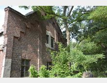 Backsteinruine in einem überwucherten, bewaldeten Gebiet mit einem kleinen, teils vermauerten Fenster und Ziegelarchitektur; umgeben von dichtem Grün und Bäumen.