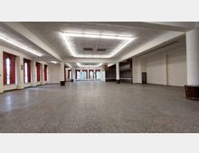 Großer leerer Veranstaltungsraum mit gefliesten Böden und einer Decke mit rechteckigen Leuchtstofflampen. An den Seiten befinden sich hohe Fenster mit roten Vorhängen, und im hinteren Bereich ist eine Theke mit holzvertäfelter Verkleidung zu sehen.