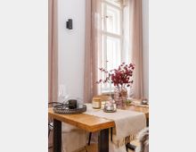 Gemütlicher Essbereich mit einem Holztisch, dekoriert mit einem cremefarbenen Stoffläufer, rot gefärbten Zweigen in einer Glasvase und weiteren Accessoires wie Kerzen und Gläsern. Im Hintergrund ein großes Fenster mit beigen Vorhängen, das natürliches Licht einfängt.