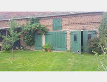 Backsteinfassade eines ländlichen Gebäudes mit grün gestrichenen Holztüren und Fensterläden. Im Vordergrund eine Wiese mit Blumen und Sträuchern, daneben eine Rankpflanze, die Teile der Wand bedeckt.